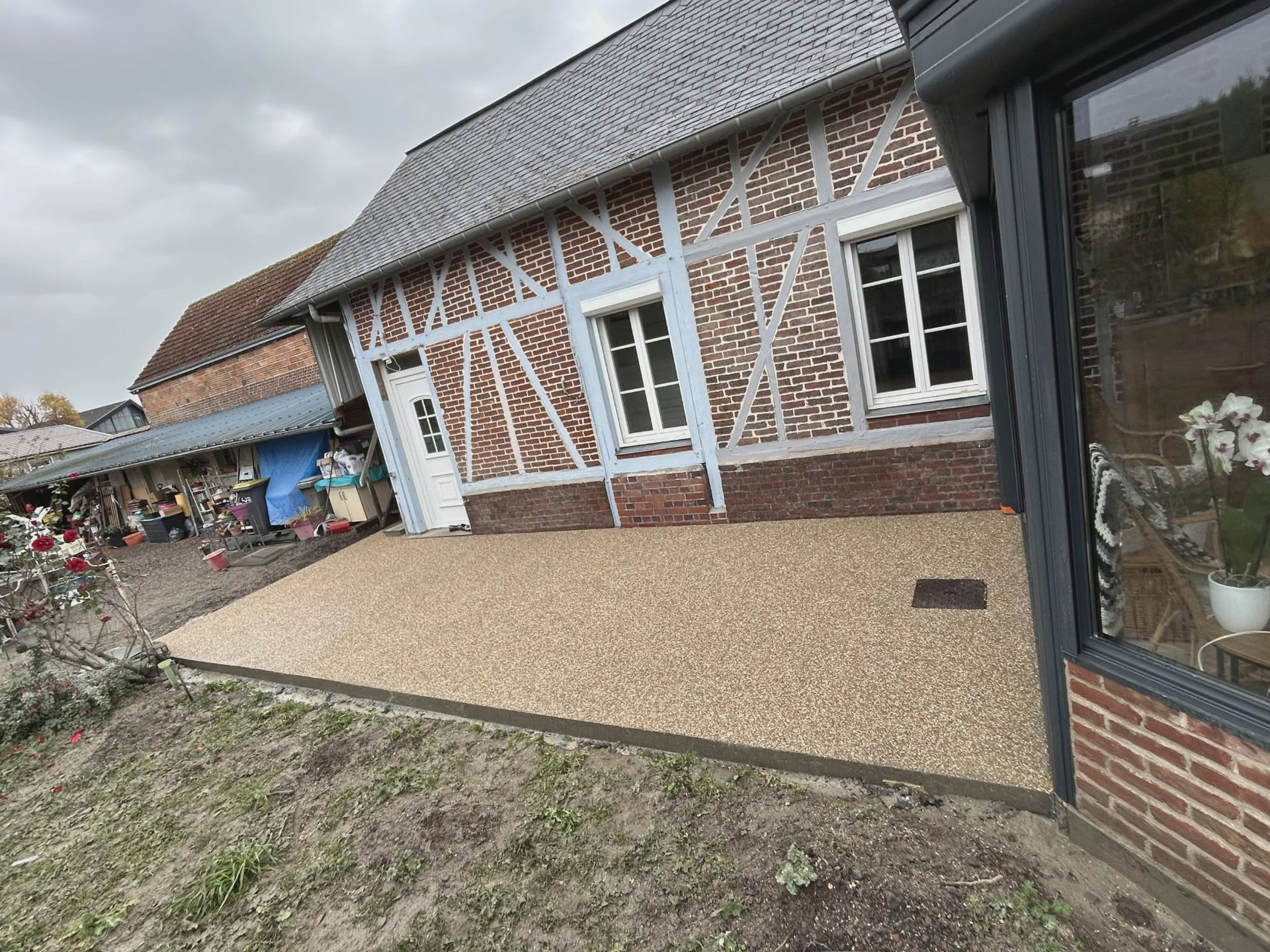 Sous un ciel nuageux, une terrasse en gravier beige recouvert de résine s'étend depuis une maison à colombages en briques.