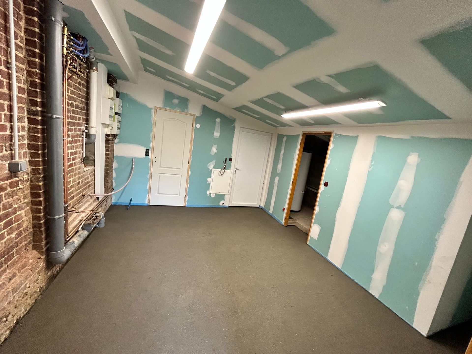 Une pièce inachevée avec un mur de briques, des panneaux de plaques de plâtre au plafond et aux murs, et un sol gris fini.