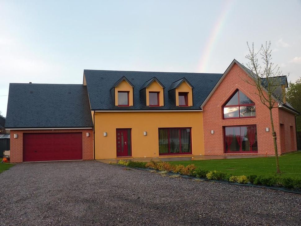 Une maison à deux étages avec un extérieur en stuc jaune, une partie de façade en briques, une porte de garage rouge et un arc-en-ciel dans le ciel.