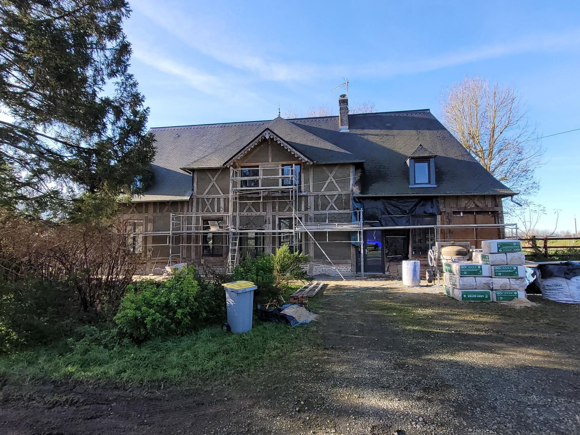 Une maison à ossature en bois de deux étages en rénovation, avec des échafaudages, entourée de verdure sous un ciel bleu azur.