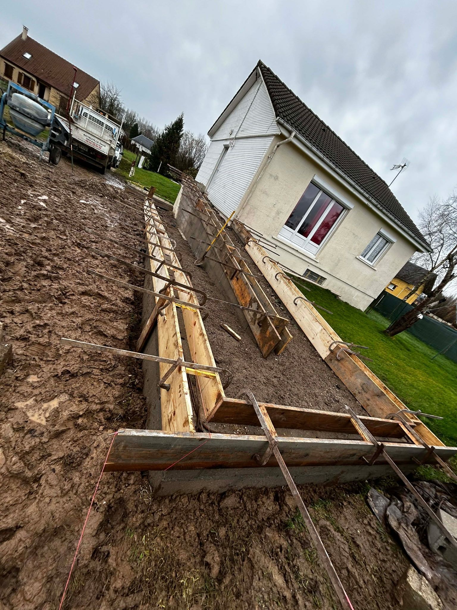 Coffrage en bois pour béton destiné à la réalisation d'une tranchée de fondation dans un jardin résidentiel boueux, près d'une maison.