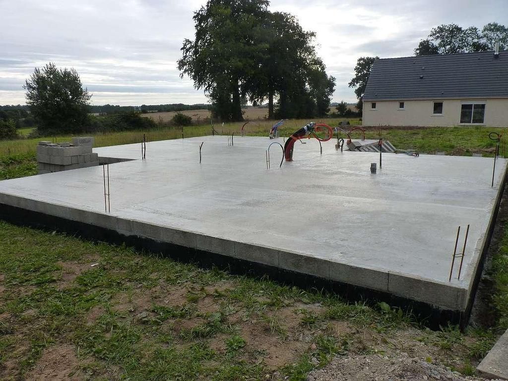 Une dalle de fondation en béton fraîchement coulée pour une maison repose sur un terrain herbeux, un bâtiment étant visible au loin.