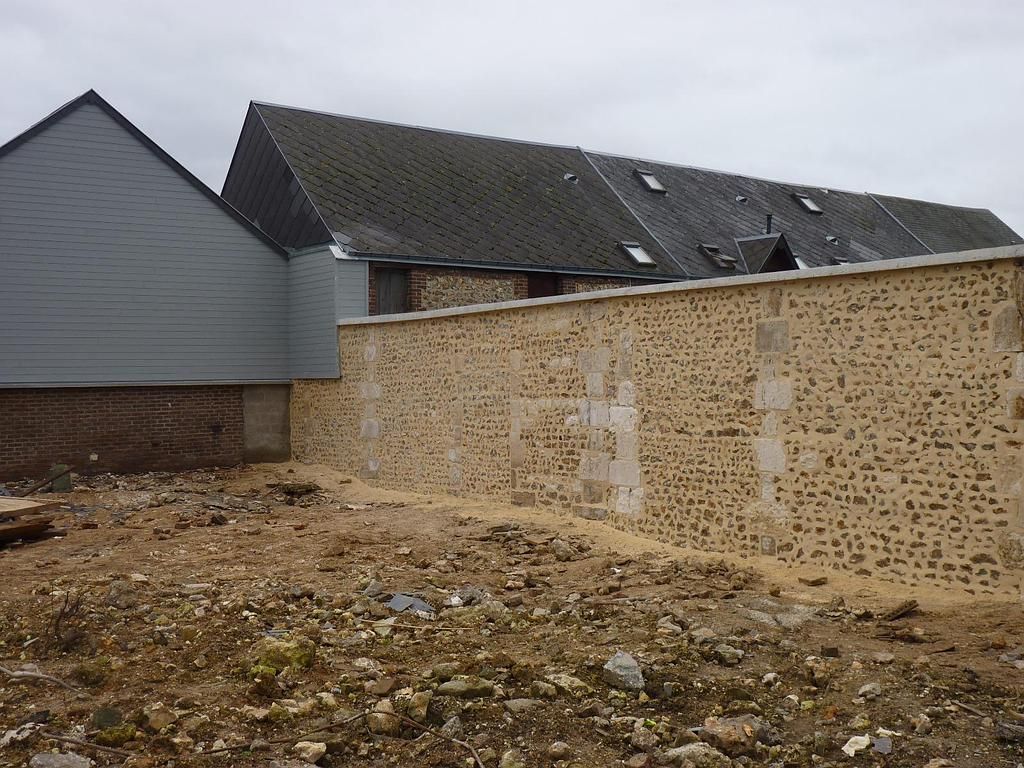 Un mur de pierre borde un terrain vague devant un bâtiment gris moderne et une maison plus grande au toit d'ardoise.