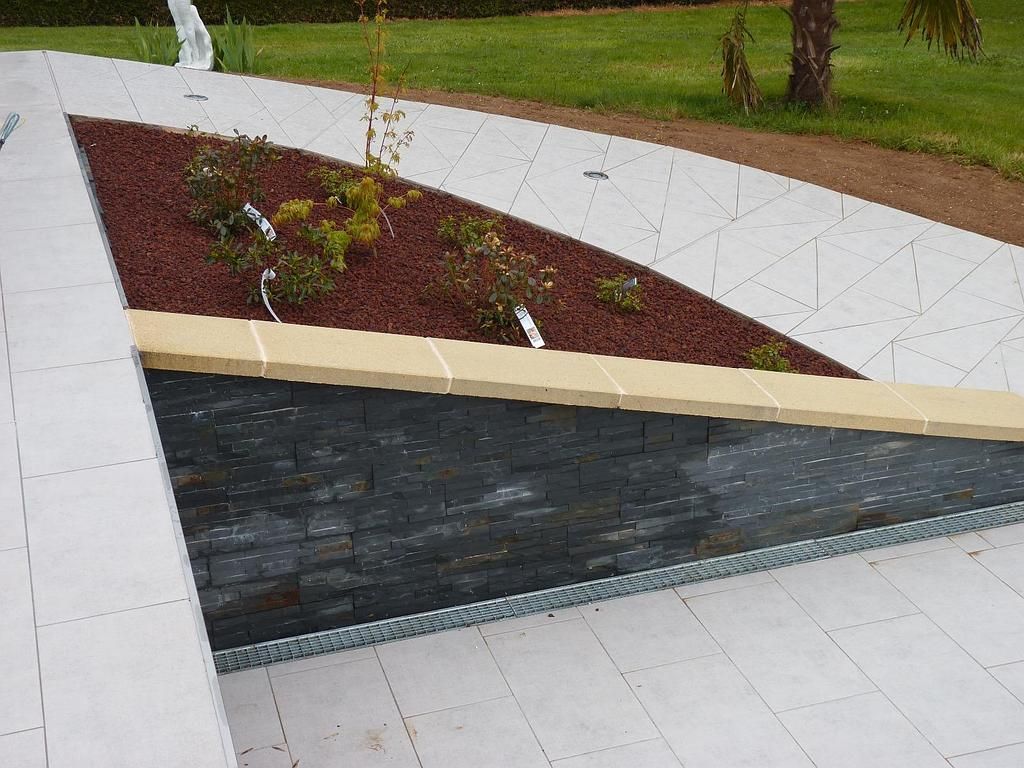 Un espace aménagé avec un mur de soutènement en pierre, du paillis rouge, de petits arbustes et des surfaces de patio carrelées de couleur claire.