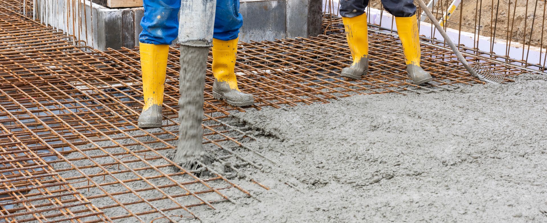 Des ouvriers du bâtiment, chaussés de bottes en caoutchouc jaunes, versent du béton frais sur un treillis métallique d'armature sur un chantier.