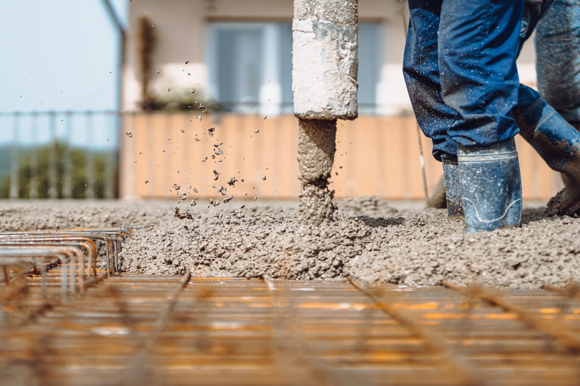 Un ouvrier en tenue de travail bleue et bottes verse du béton frais sur une grille d'armature en acier sur un chantier de construction.