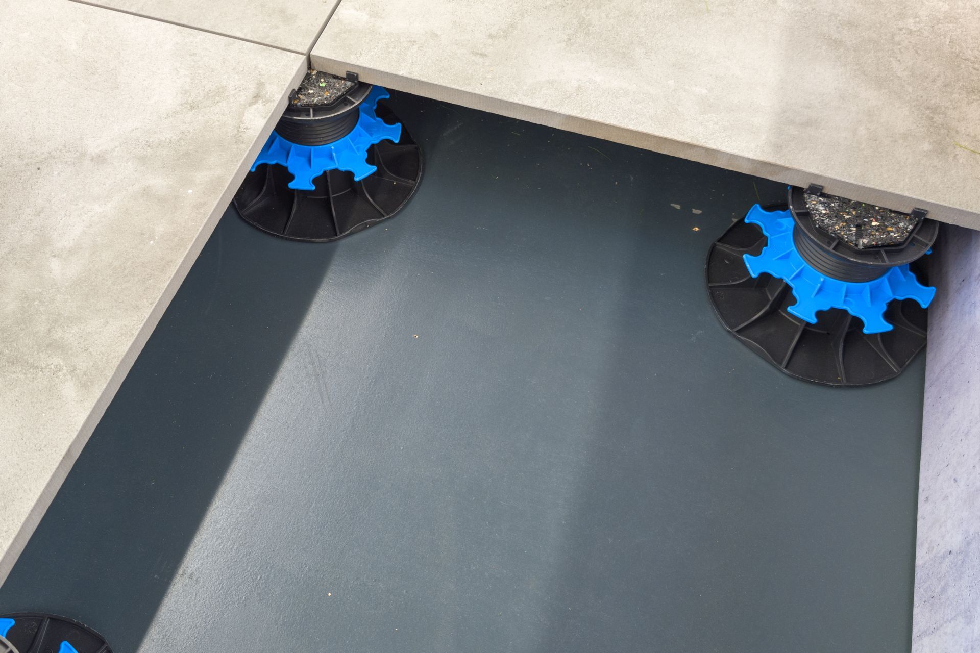 Dalles de béton installées sur des plots réglables en plastique noir et bleu, le tout sur une surface sombre.