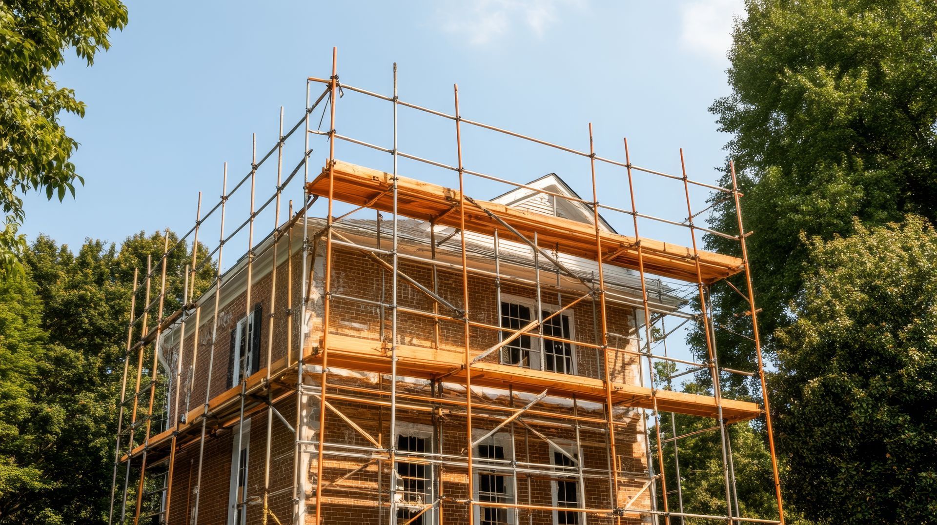 Une maison en briques en cours de rénovation, entourée d'échafaudages métalliques, se détachant sur un fond d'arbres verts et de ciel bleu.