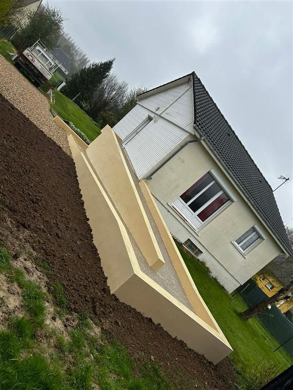 Un nouveau mur de soutènement clair borde une parcelle de terre et d'herbe fraîchement semée, jouxtant une maison au toit sombre.