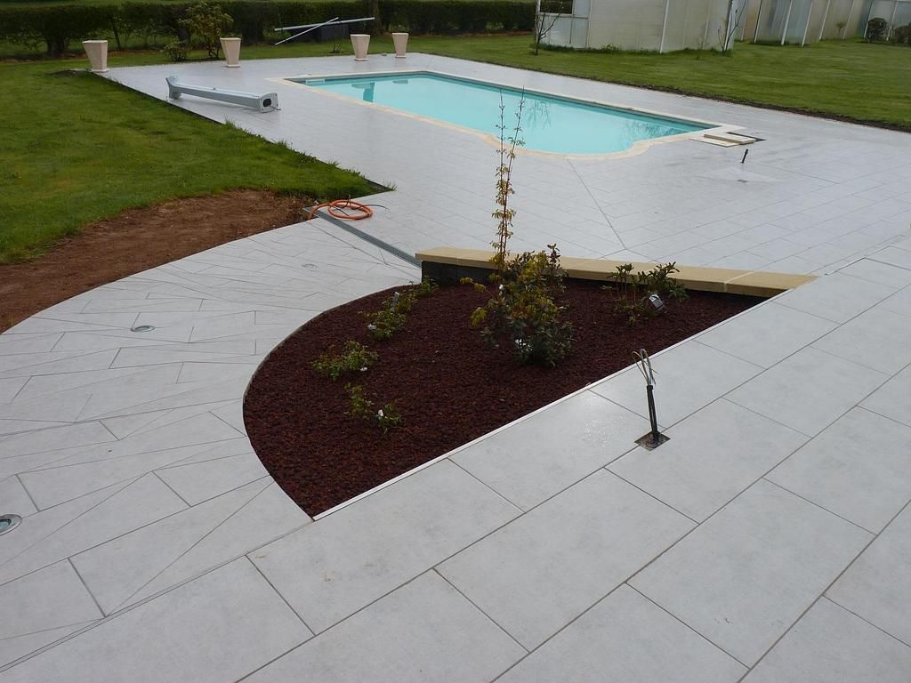 Une piscine rectangulaire entourée d'une terrasse pavée gris clair avec un parterre de fleurs incurvé recouvert de paillis foncé.