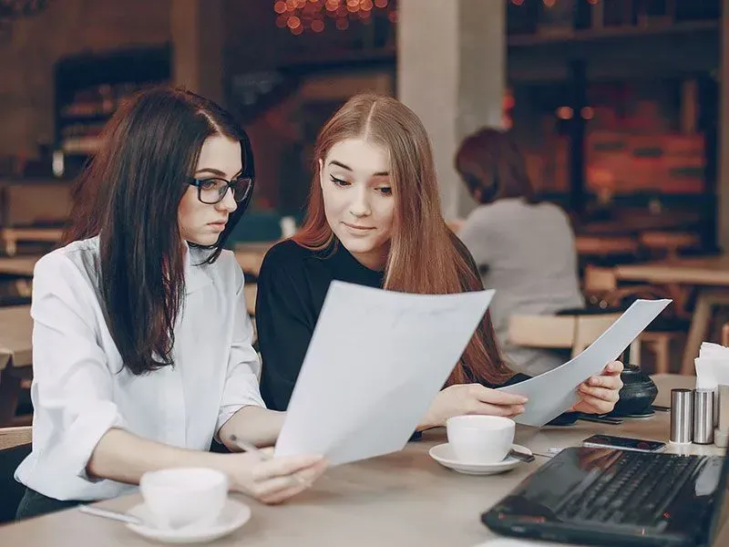 Dos mujeres examinan papeles en un café, con expresiones concentradas; sobre la mesa hay tazas de café