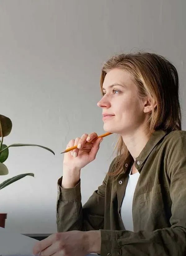 Mujer con camisa verde, sosteniendo un lápiz junto a su barbilla, con expresión pensativa cerca de una planta en maceta.