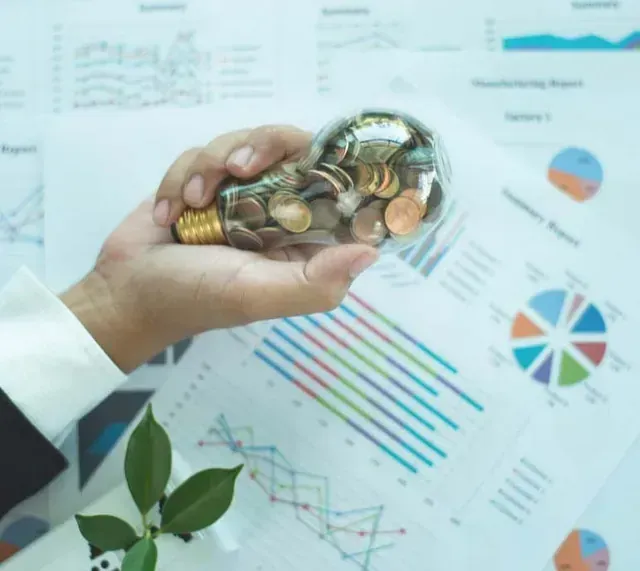 Una mano sostiene una bombilla llena de monedas, con gráficos financieros y una pequeña planta de fondo.