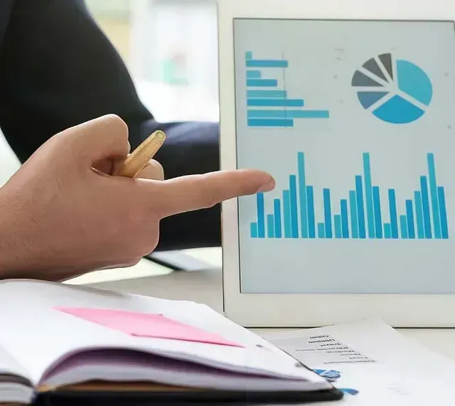 Persona señalando la pantalla de una tableta que muestra gráficos y tablas financieras; cuaderno y bolígrafo cerca.