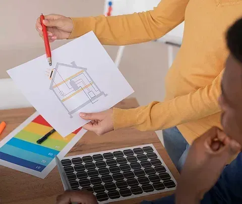 Persona señalando el plano de una casa con un panel solar y una tabla de colores sobre la mesa.