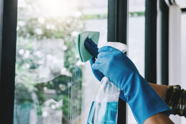 Eine Hand mit blauen Handschuhen reinigt ein Fenster mit einer Sprühflasche und einem Tuch.