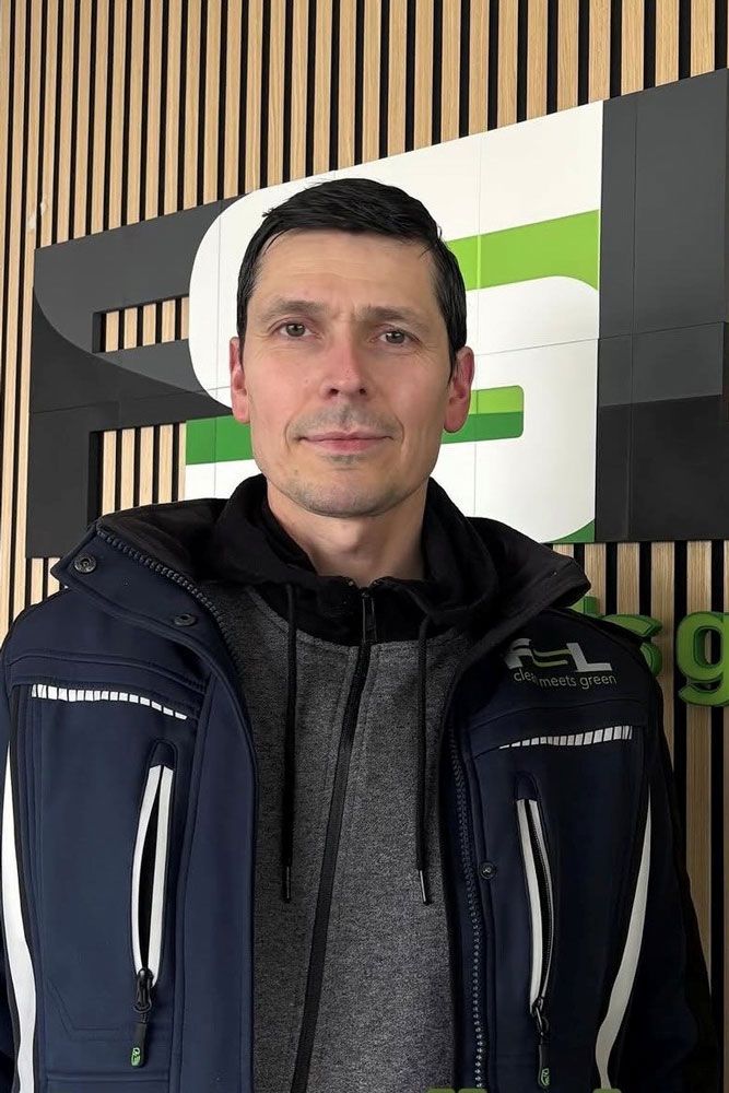 Ein Mann steht in einem Innenraum vor einem grün-weißen Schild und trägt eine dunkle Jacke und einen Kapuzenpullover.