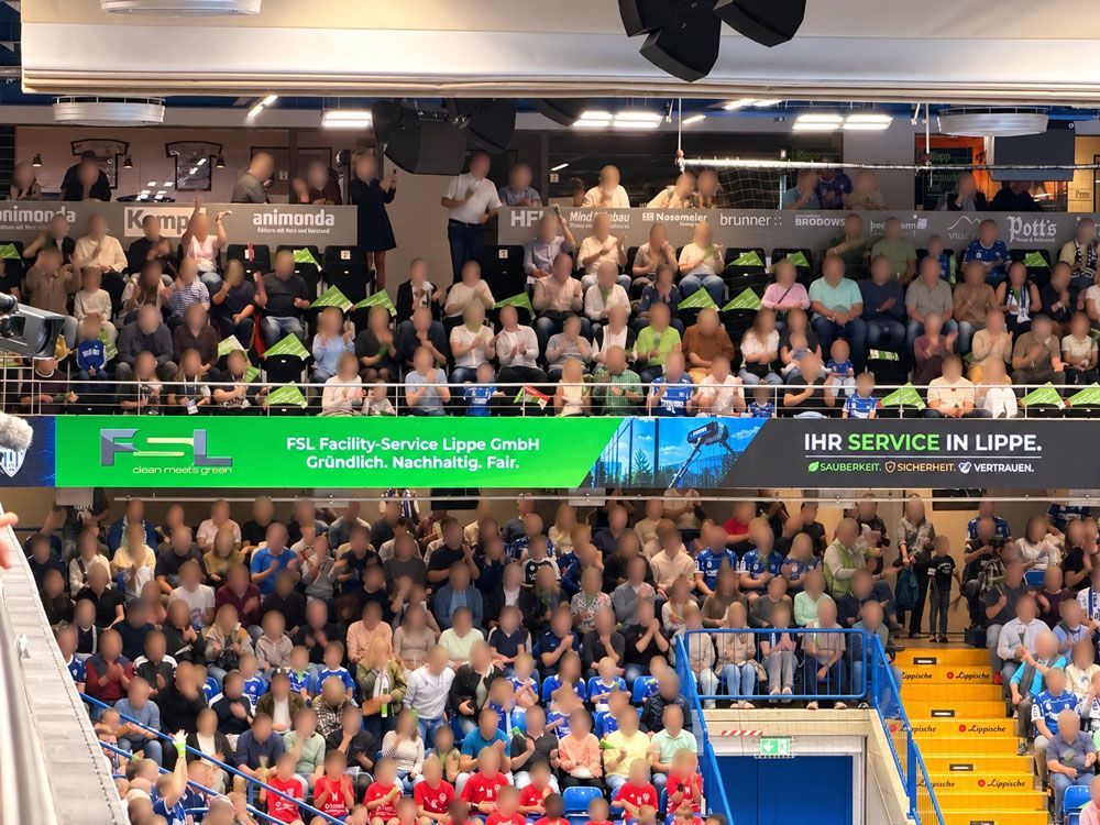 In einem hellen Saal verfolgt das Publikum eine Live-Sportübertragung auf einer Großleinwand.