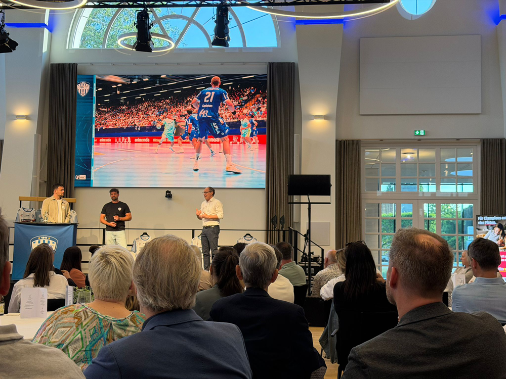 In einem hellen Saal verfolgt das Publikum eine Live-Sportübertragung auf einer Großleinwand.