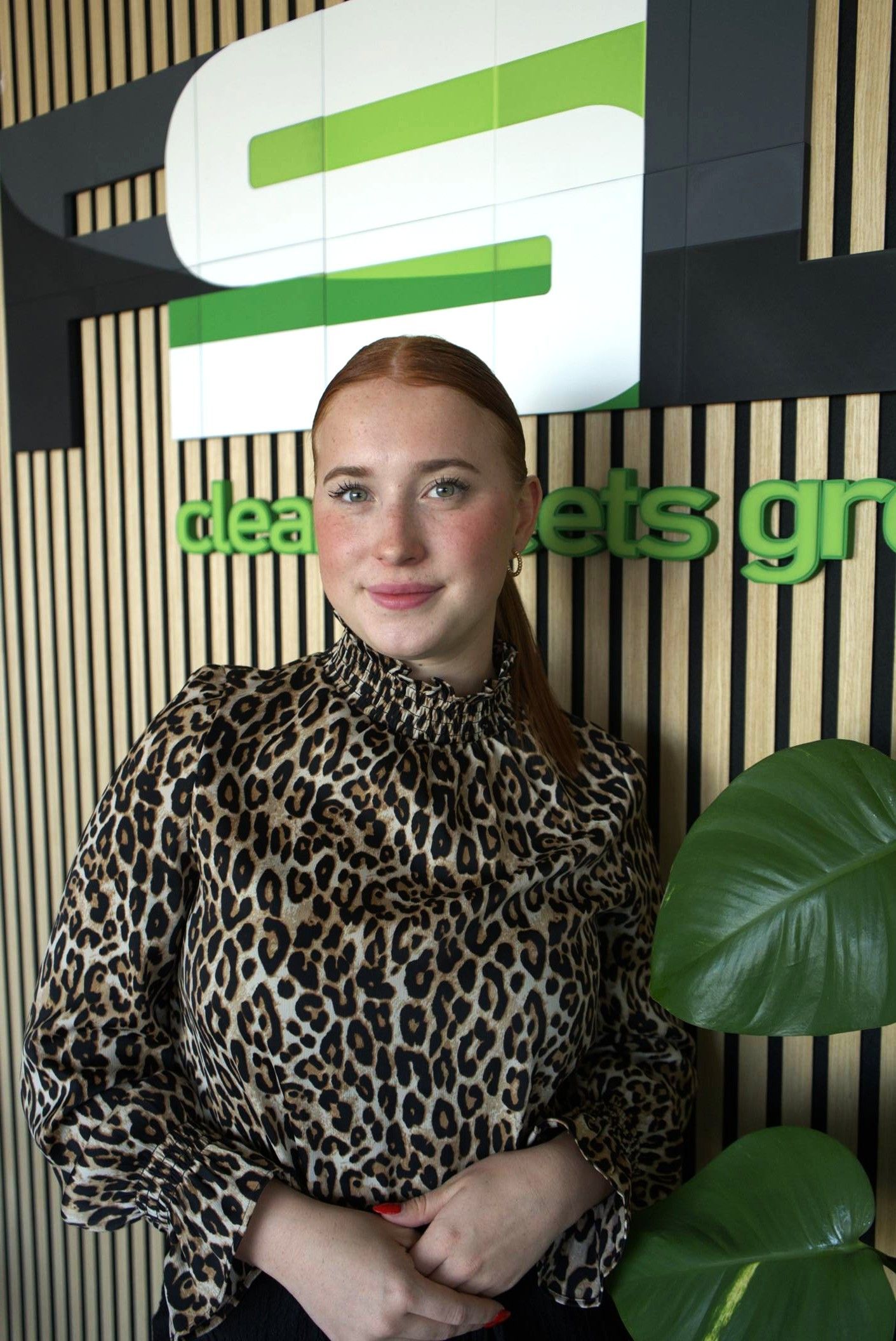 Eine Frau in einem Leopardenmuster-Top steht vor einem grün-schwarzen Büroschild; in der Nähe befinden sich Topfpflanzen.