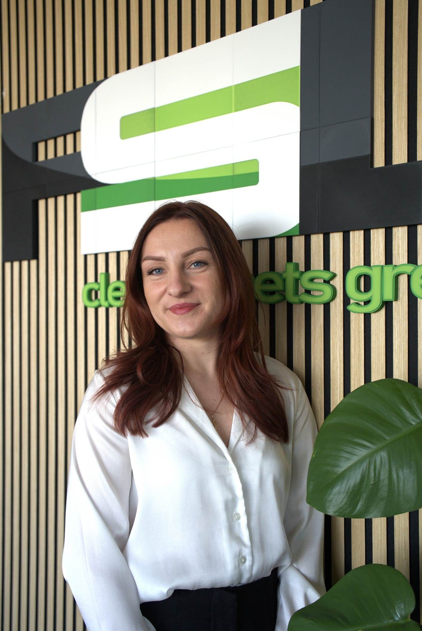 Eine Frau mit weißer Bluse steht in der Lobby eines Büros vor einem grünen Logo und einer Wand aus Holzlatten.