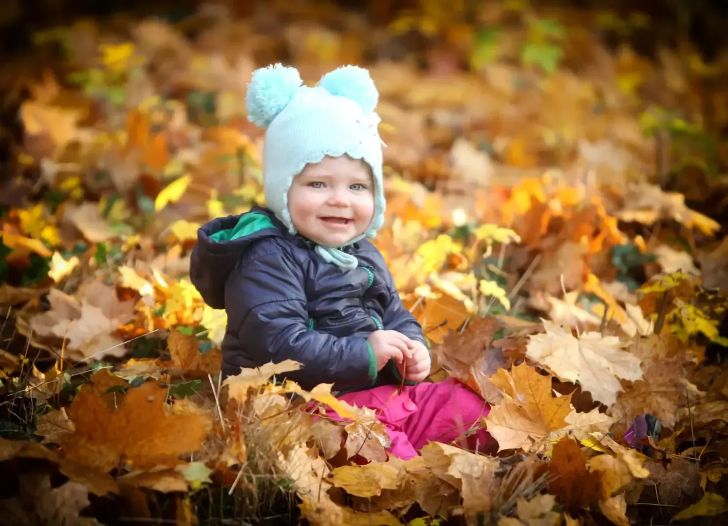 Ein lächelndes Kind mit hellblauer Bommelmütze und dunkler Jacke sitzt inmitten eines Haufens bunter Herbstblätter.