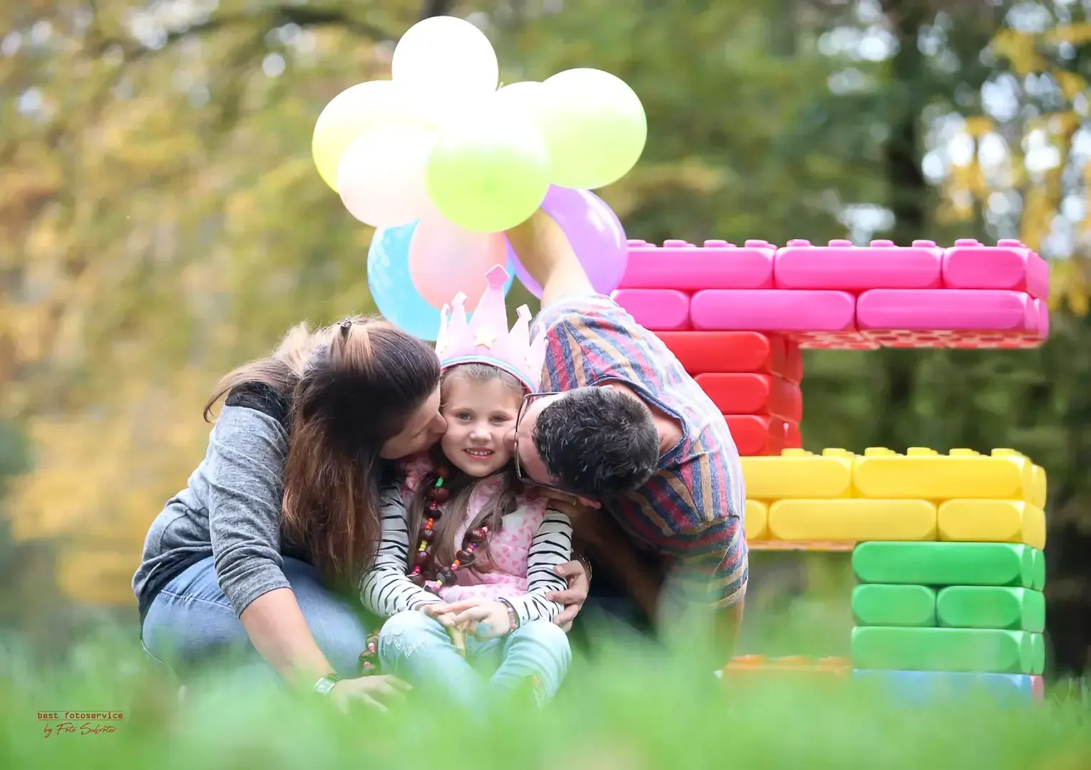 Die Eltern küssen ihre Tochter, die draußen neben einer großen Fünf aus Bausteinen und bunten Luftballons eine Krone trägt.
