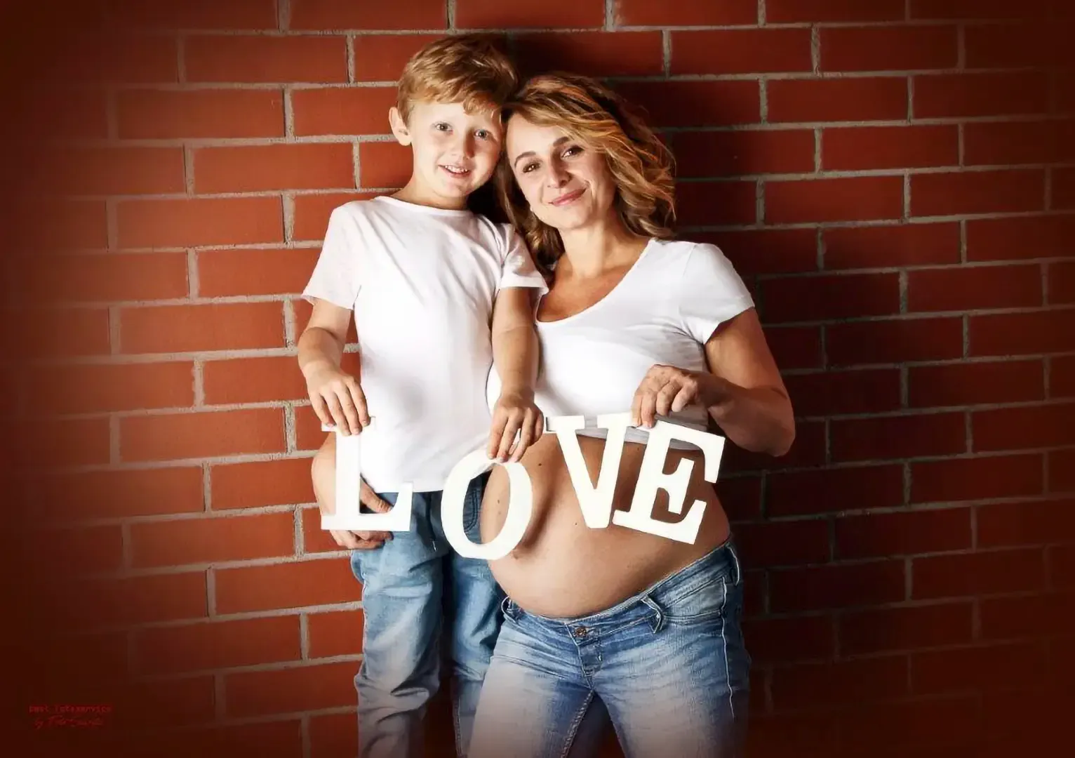 Eine schwangere Frau und ein Kind stehen an einer Backsteinmauer und halten große weiße Buchstaben, die das Wort „LOVE“ bilden, über ihren Bäuchen.