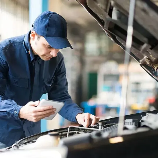 Un mecánico con uniforme y gorra azules inspecciona el motor abierto de un coche mientras mira una tableta