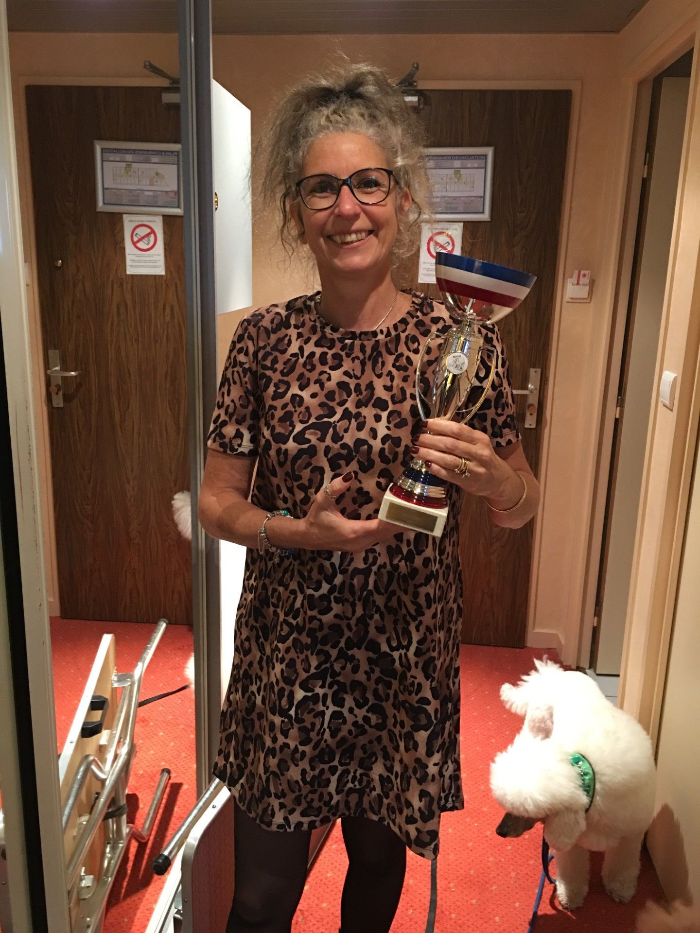 Une femme tenant un trophée se tient dans un couloir avec un chien blanc. 