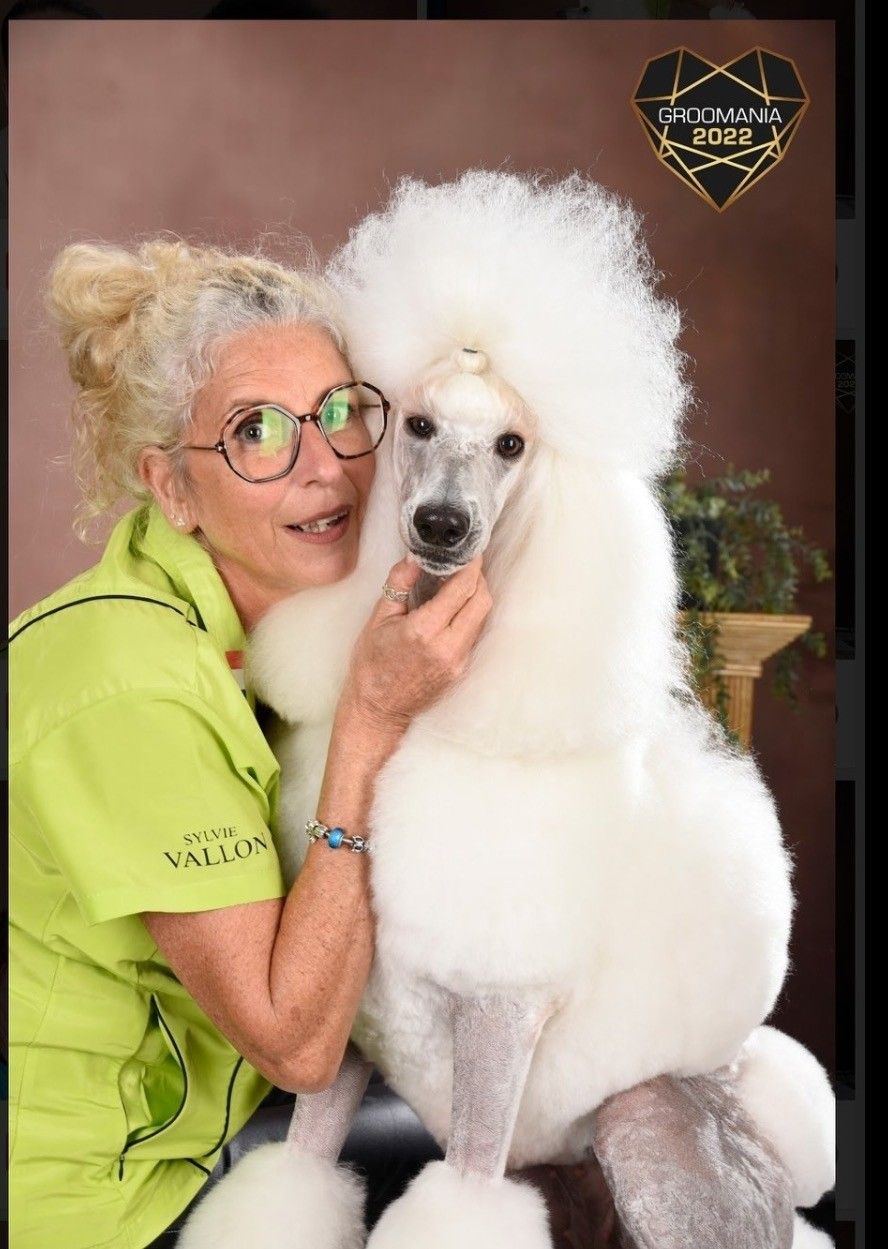 Une femme à lunettes enlace un caniche blanc ; toutes deux sourient. En arrière-plan : mur marron, petite plante.