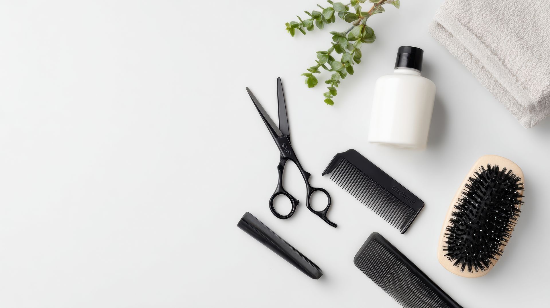 Outils de coiffure sur une surface blanche : ciseaux, peignes, brosse, lotion, serviette et brin de feuilles vertes.