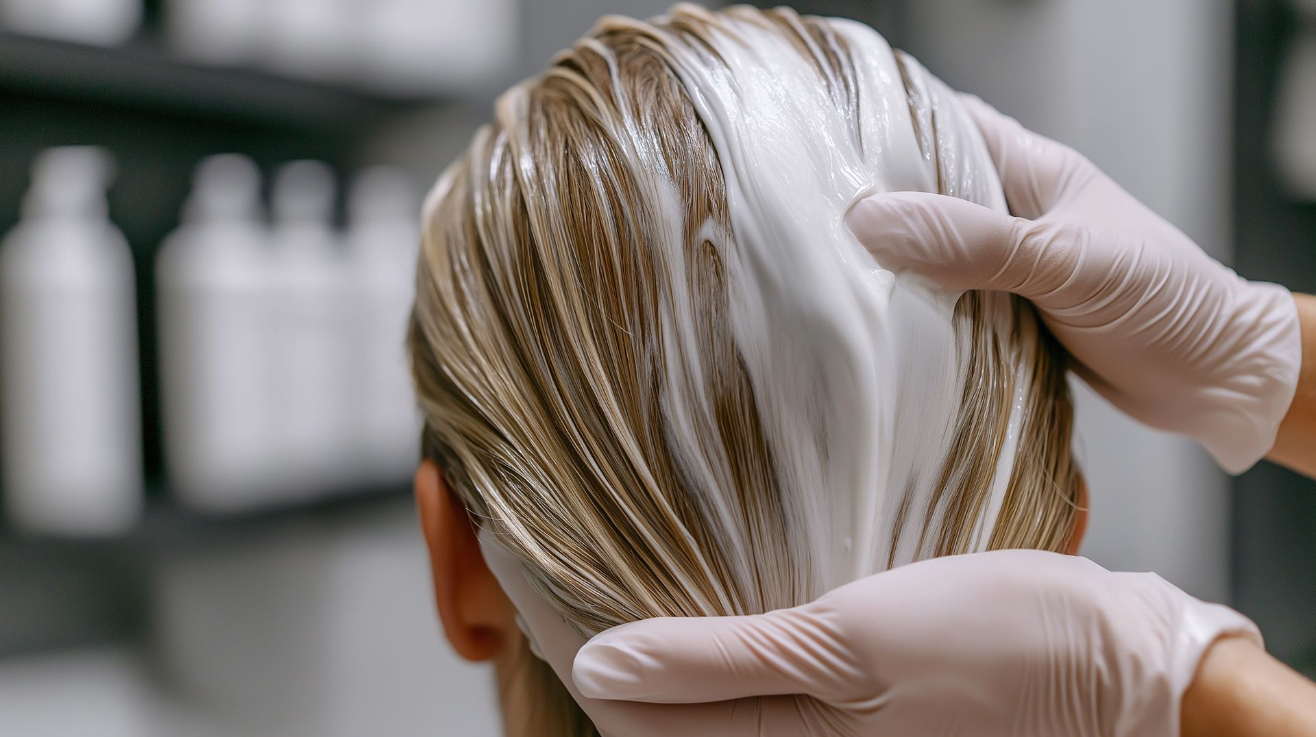Des mains gantées appliquent un soin capillaire sur des cheveux blonds dans un salon.