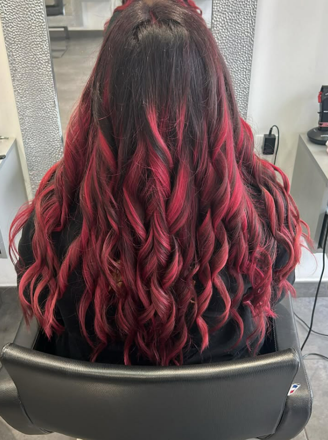 Femme aux longs cheveux ondulés, aux racines foncées et aux pointes rouge vif, assise sur un fauteuil de salon.