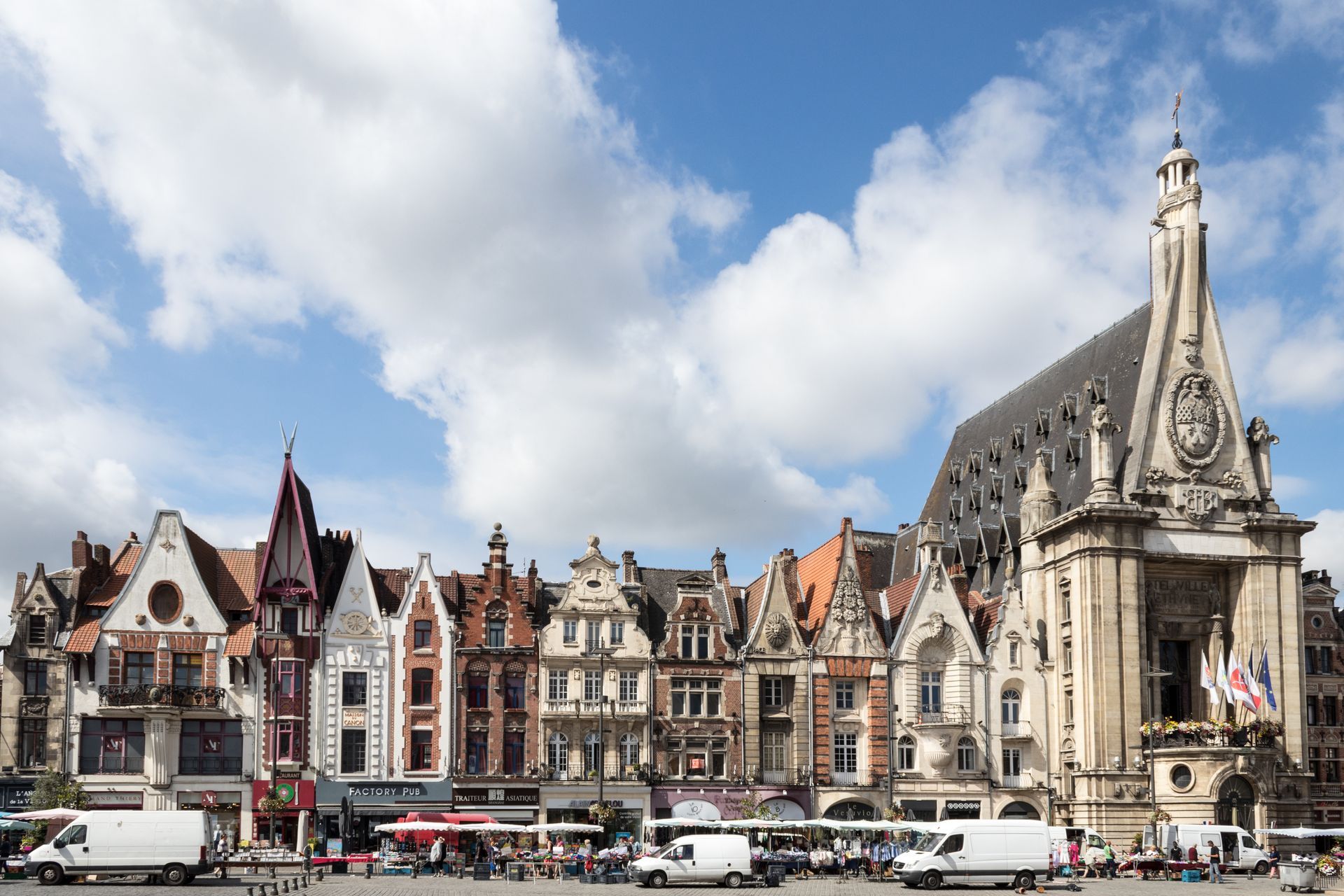 Des bâtiments aux façades ornées bordent la grand-place de Béthune sous un ciel bleu parsemé de nuages ​​cotonneux.