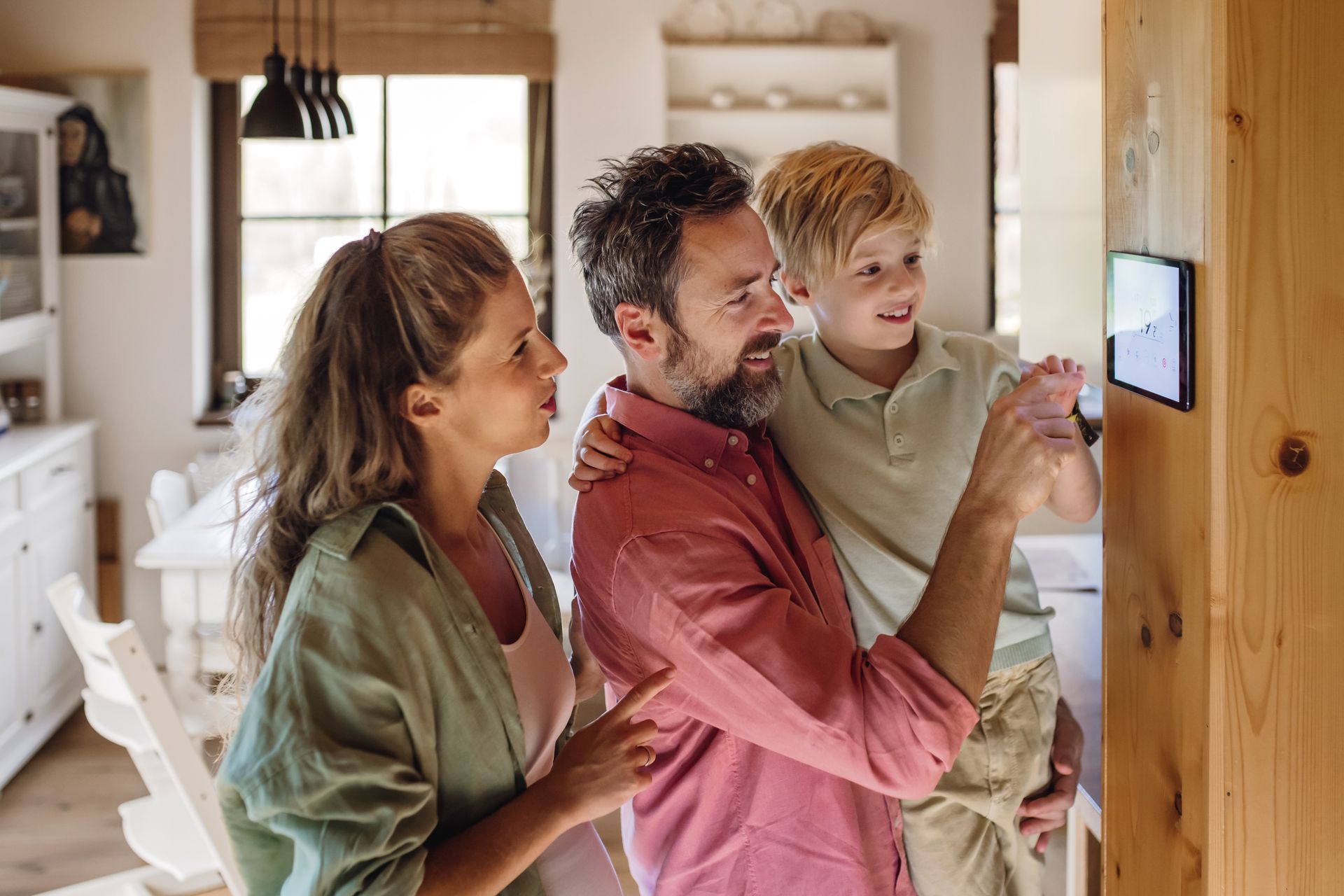 Une famille regarde une tablette fixée au mur en bois, tout en souriant. À l'intérieur, dans la cuisine.