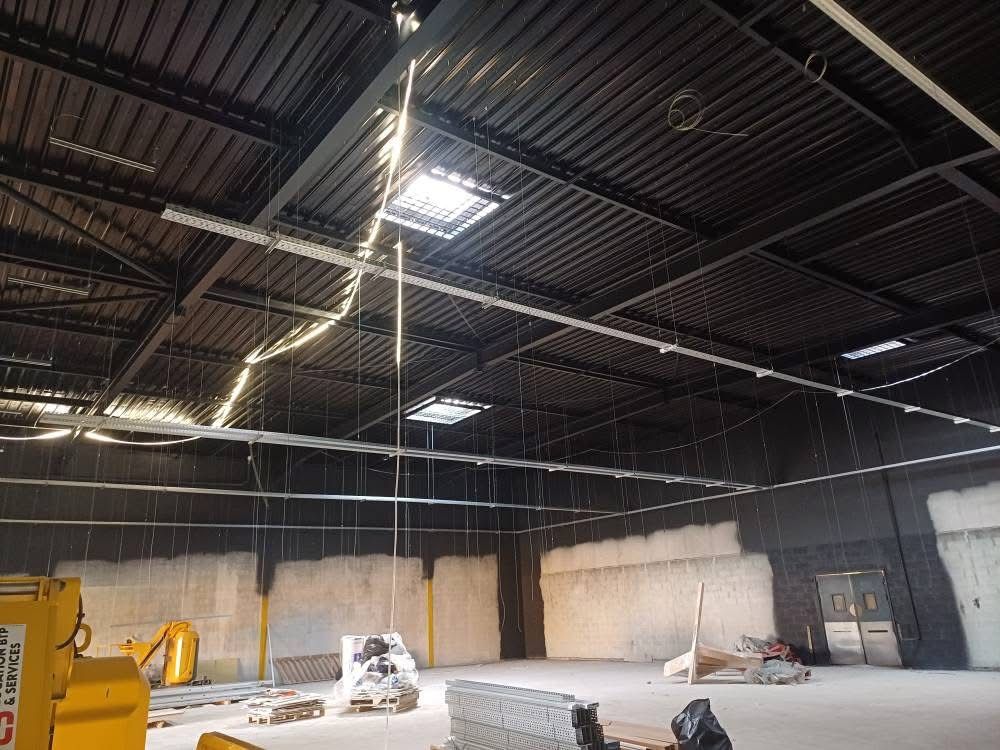 Vue intérieure d'un chantier de construction d'entrepôt avec hauts plafonds, poutres apparentes et murs inachevés ; éclairage et équipements visibles.