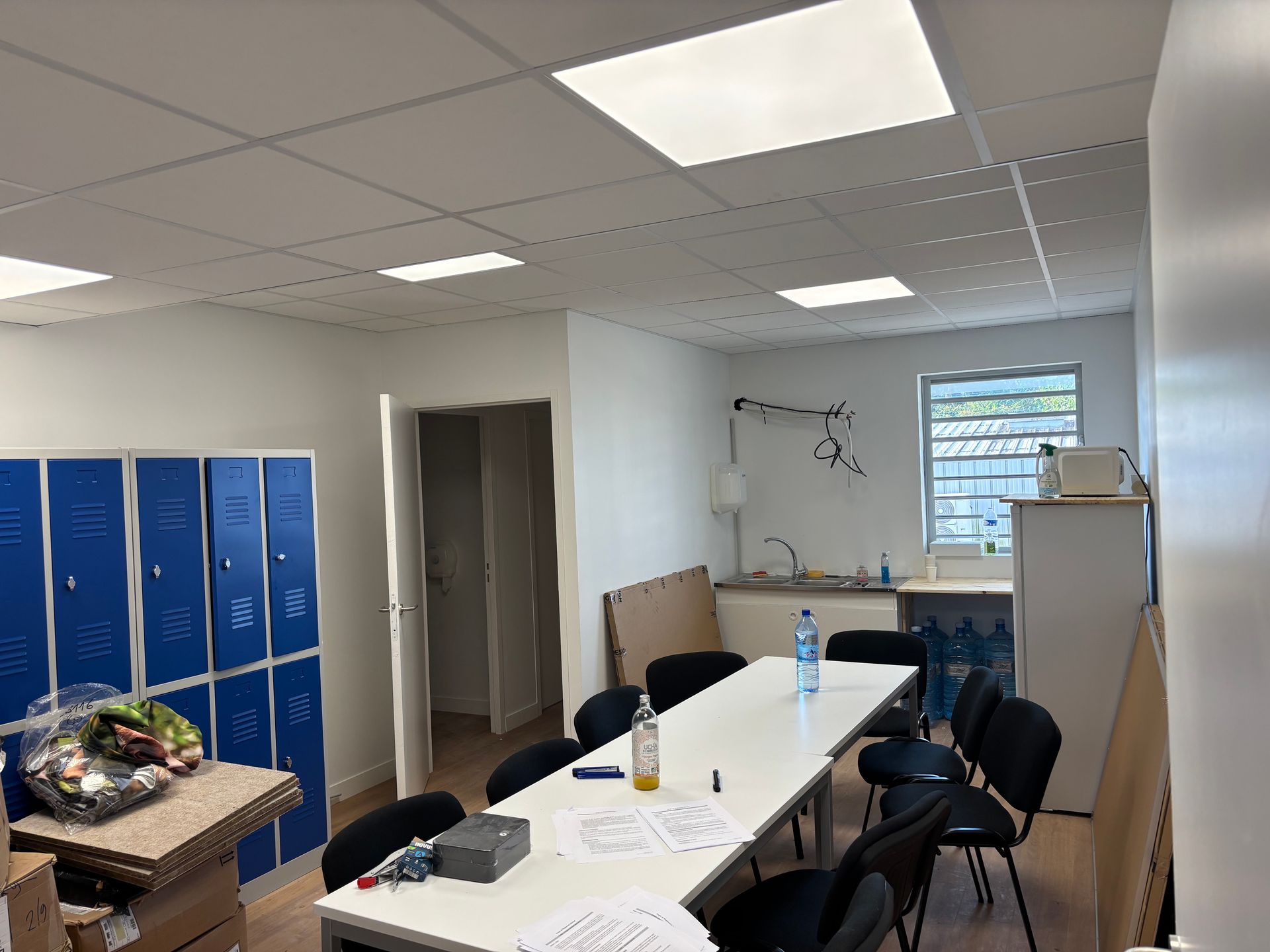 Un bureau vide avec des casiers, une table, un lavabo et une porte ouverte. Murs blancs, casiers bleus et chaises noires.