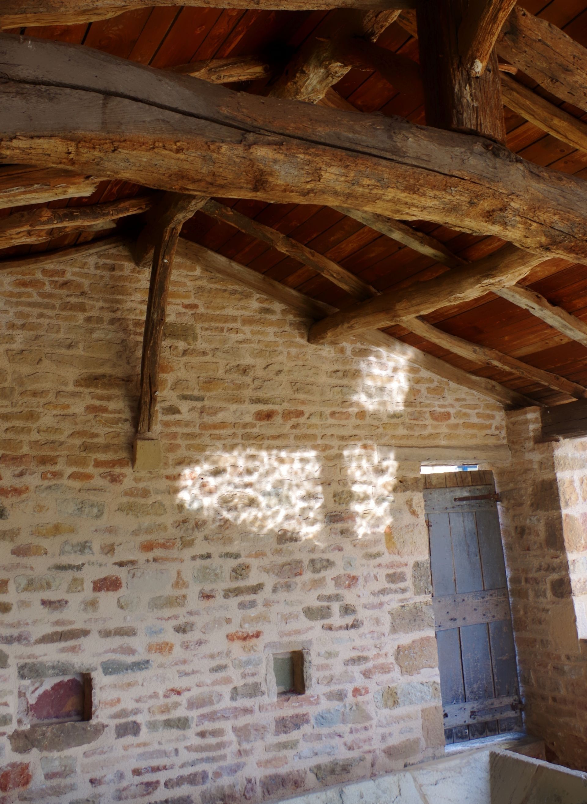 Pièce rustique en pierre avec poutres apparentes et une porte, éclairée par la lumière du soleil à travers une petite fenêtre