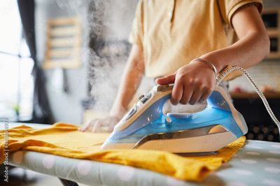 Eine Person bügelt ein gelbes Hemd auf einem Bügelbrett mit einem blauen Dampfbügeleisen.