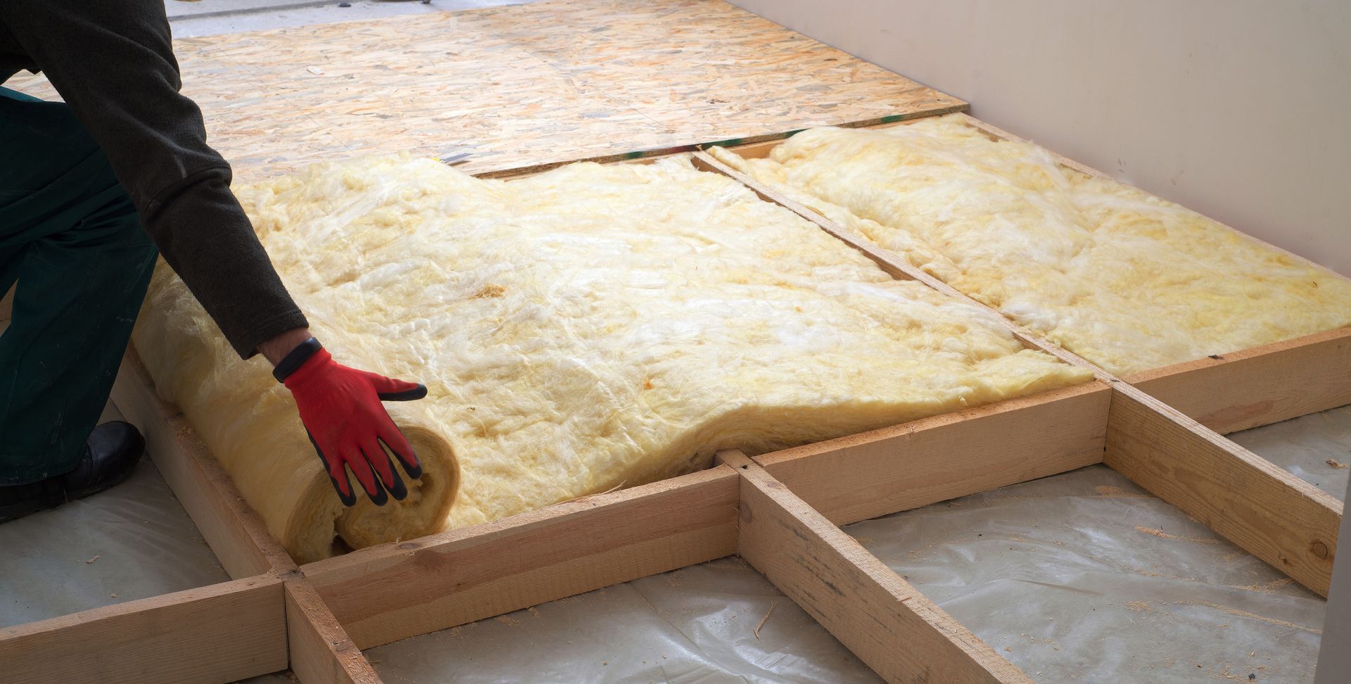 Une personne déroule de l'isolant entre les solives d'un plancher en bois sur une surface blanche.