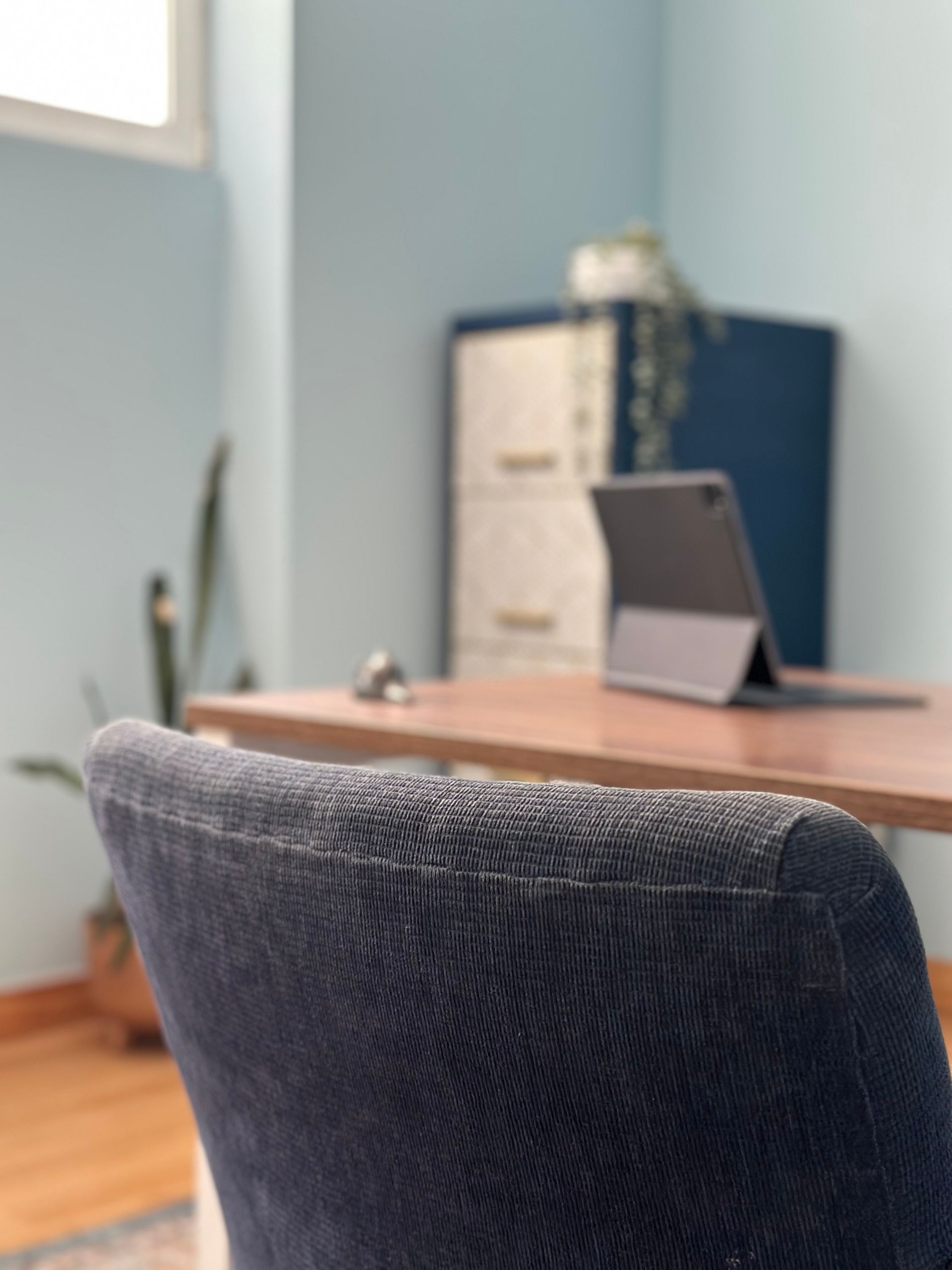 Acogedora oficina en casa con un escritorio de madera, un portátil y una silla azul oscuro junto a una ventana luminosa.