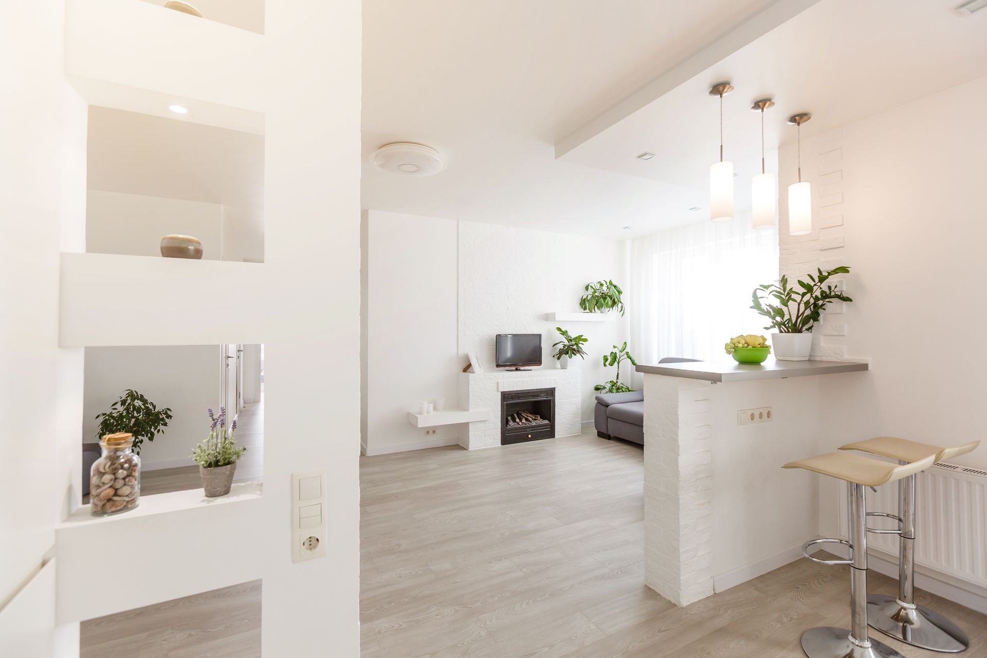 Intérieur d'appartement moderne et blanc lumineux ; bar avec tabourets, plantes, télévision, cheminée et lumière naturelle.
