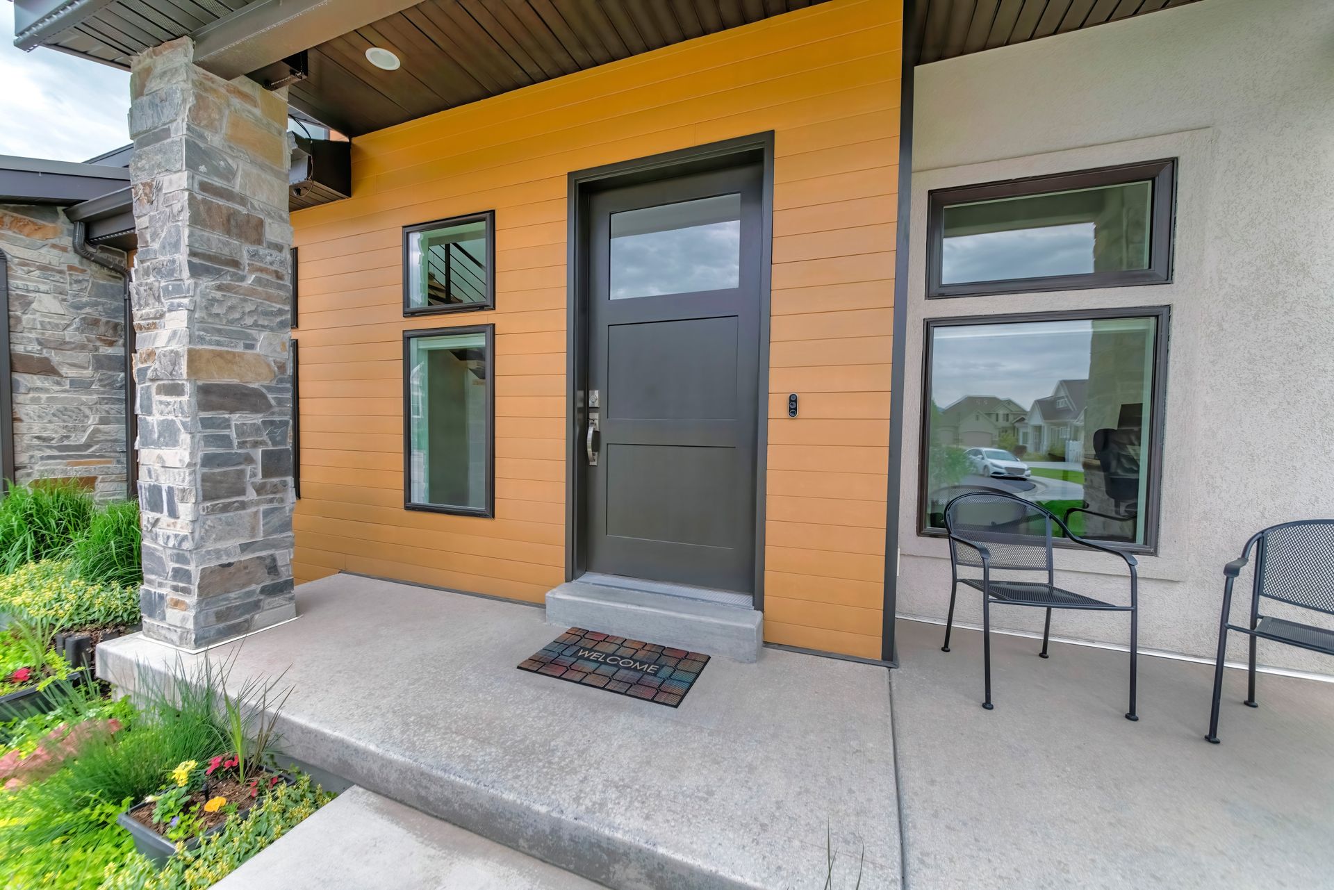 Entrée de maison moderne avec porte sombre, pilier en pierre et petites fenêtres.