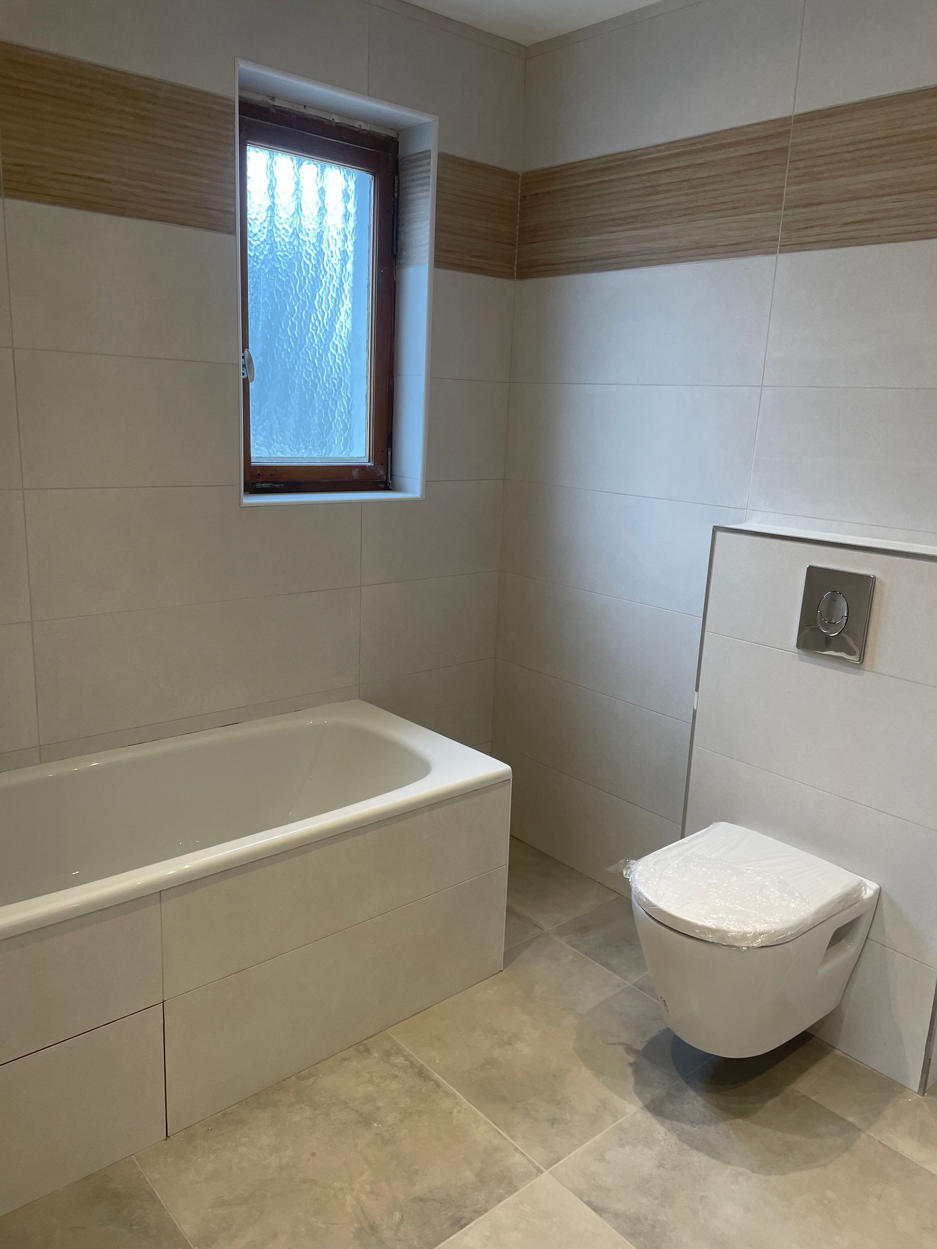 Salle de bains avec baignoire blanche, toilettes, fenêtre et carrelage beige aux murs et au sol.