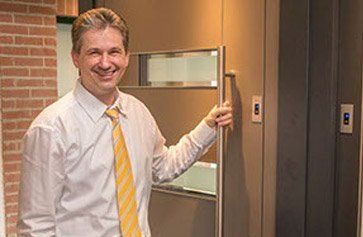 Ein Mann in einem weißen Hemd und einer gelben Krawatte steht vor einem Aufzug.