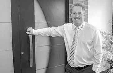 Mann in weißem Hemd und Krawatte, lächelt, während er eine Türklinke hält; Außeneinstellung.