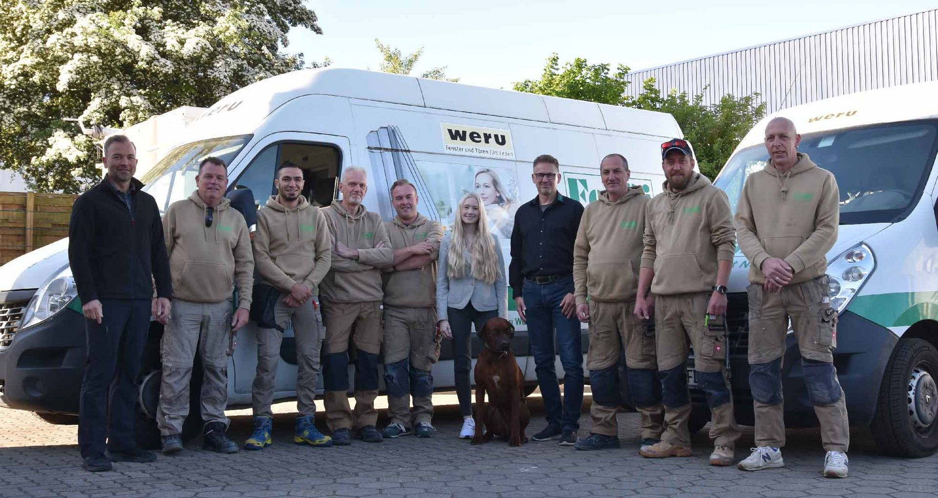 Eine Gruppe von Menschen vor zwei weißen Lieferwagen mit Logos. Einige tragen Uniformen und einen Hund.