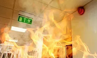 Pasillo envuelto en llamas con alarma de incendios y señal de salida visibles.