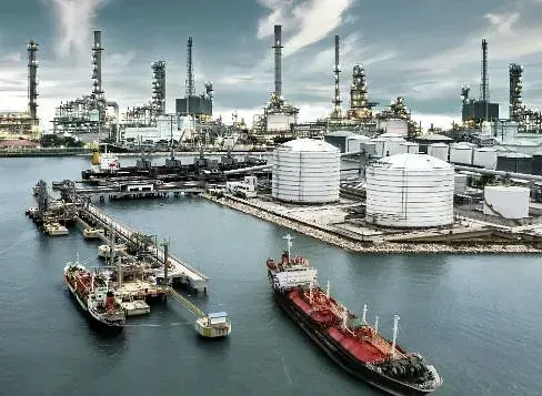 Refinería de petróleo con grandes tanques, tuberías y barcos de carga atracados en un puerto, bajo un cielo nublado.
